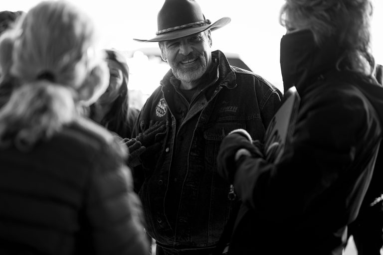 Black and white photograph of Pastor Troy Brewer in a hat and denim jacket, talking with people at the food bank.